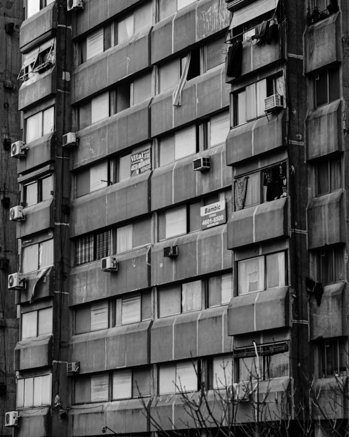 Black and white image of a city apartment building with visible air conditioners and window details.